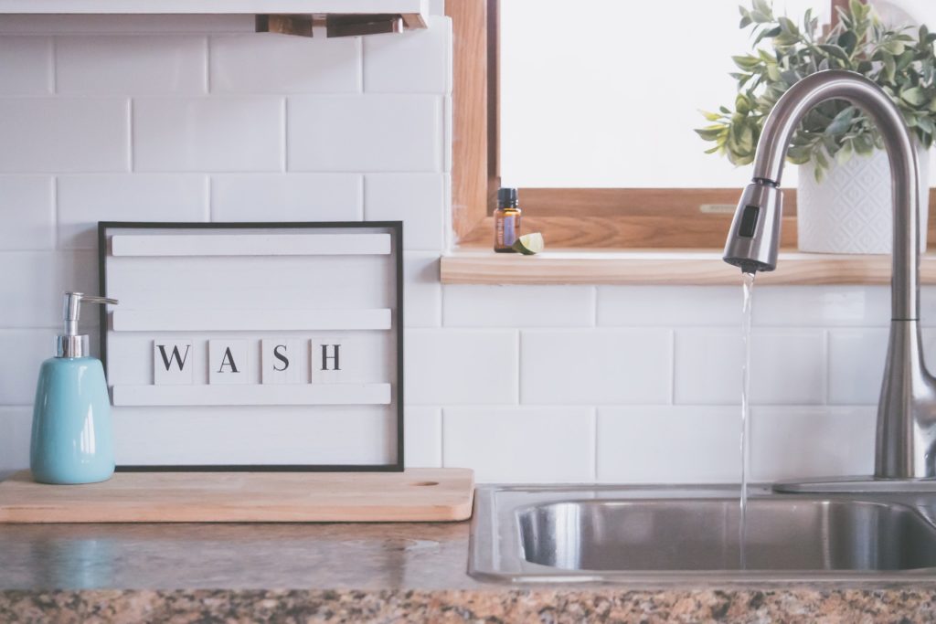 Board with word WASH placed near soap dispenser on counter in bathroom with water streaming from open faucet