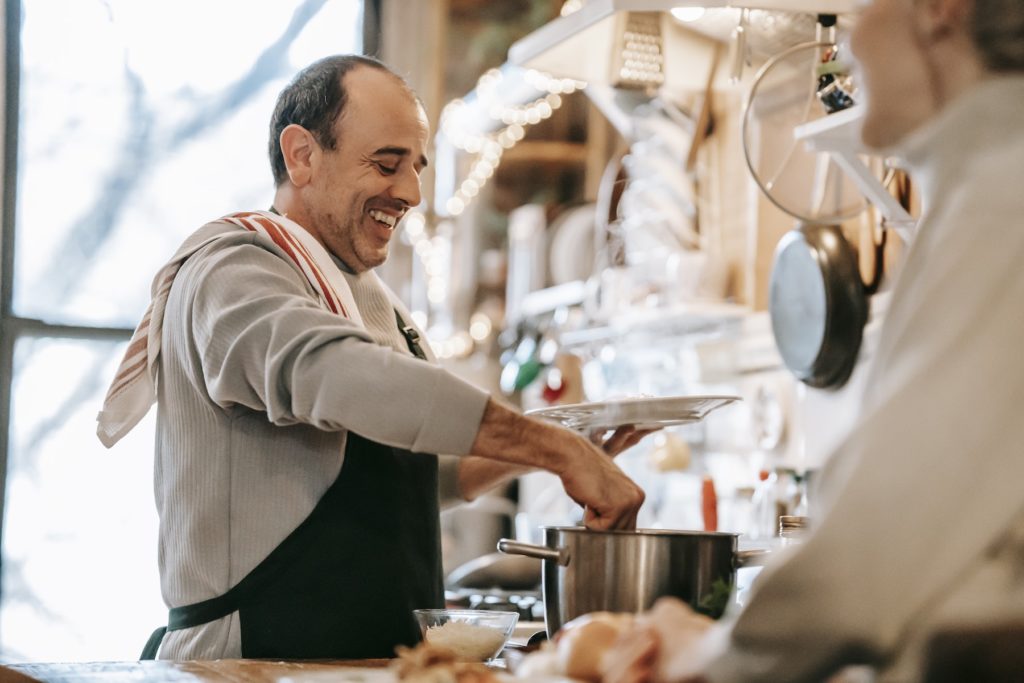 Positive ethnic man smiling and serving homemade dish for unrecognizable wife